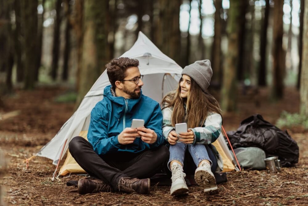 cheerful-young-european-man-and-female-tourists-in-jackets-resting-in-forest-near-tent-typing-on-e1733326197770